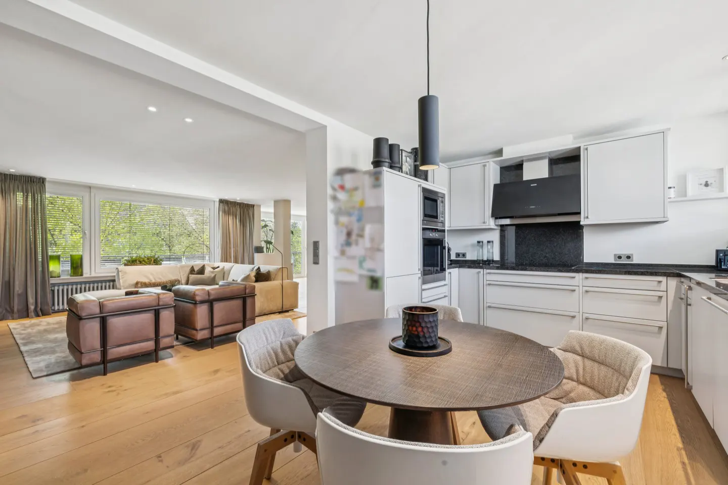 Open-concept living space with a round dining table, white chairs, and a modern kitchen with white cabinets and black countertops. A living room is visible in the background.