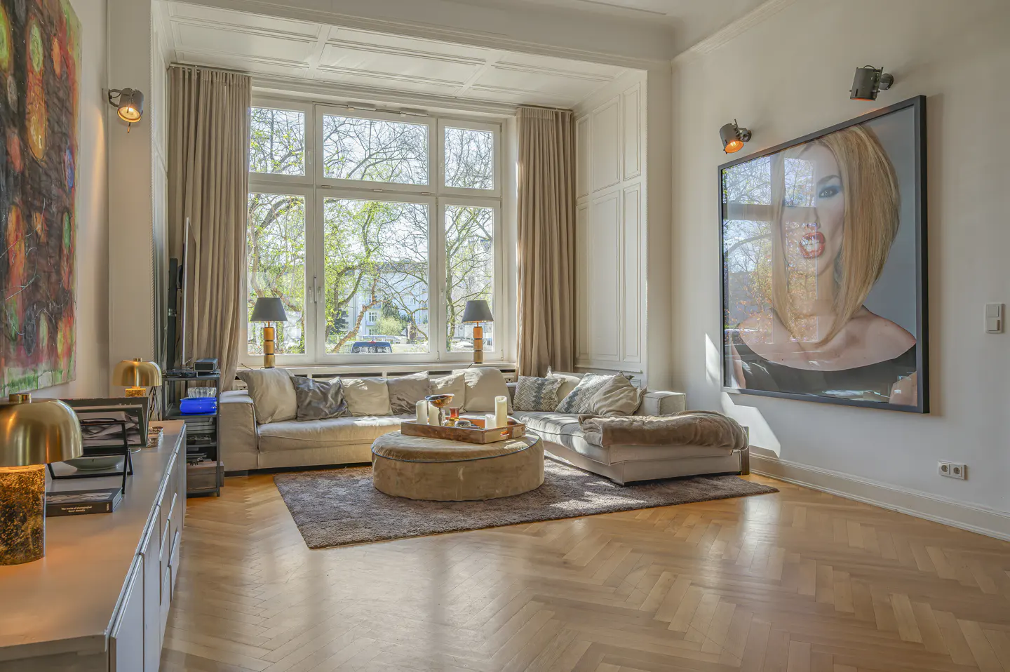 Bright living room with herringbone wood floors, large window, beige sofas, and a framed portrait of a woman.