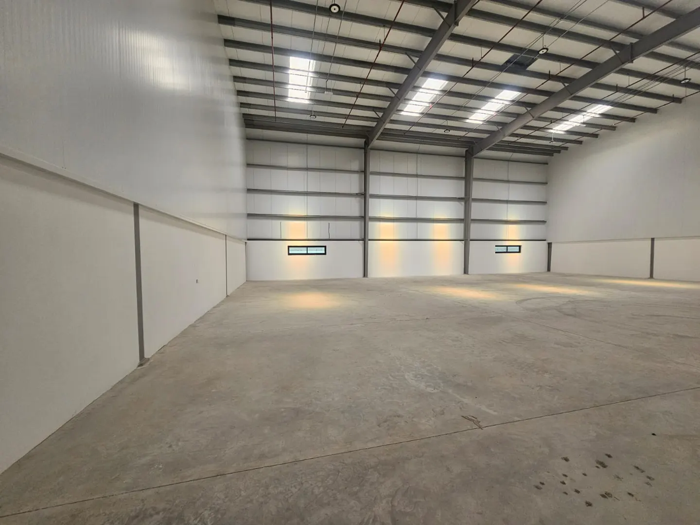 Empty warehouse interior with concrete floor, white walls, and a high ceiling with metal beams and skylights.