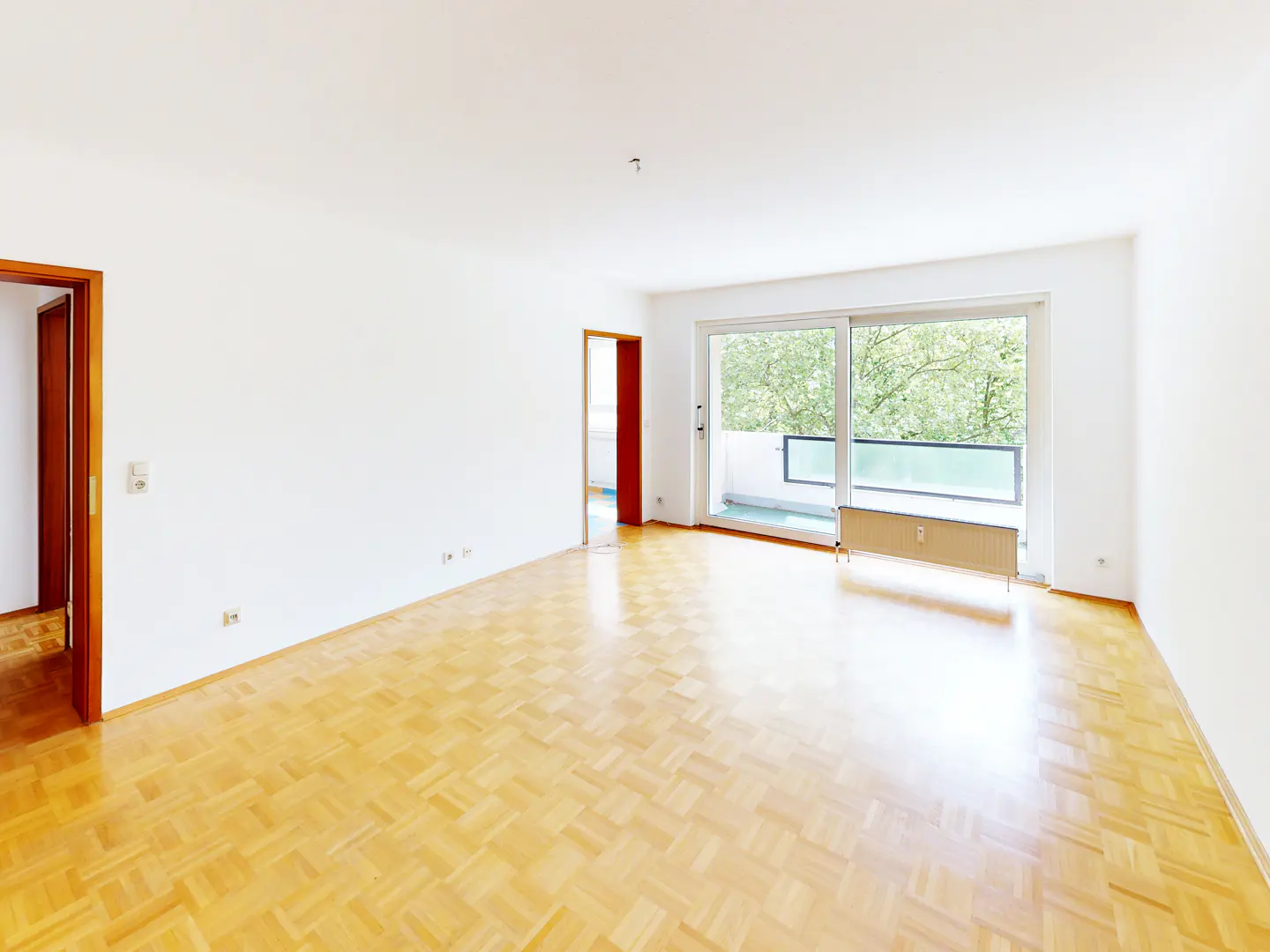 Bright, empty room with white walls, light wood parquet flooring, and a large sliding glass door leading to a balcony.