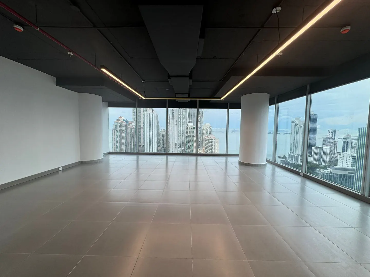 Empty office space with gray tile floor, white walls, and a black ceiling with linear lighting. Large windows offer a city view.