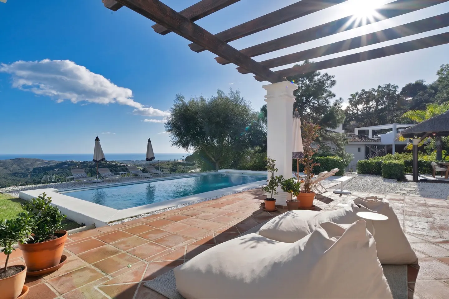 Outdoor patio with pool overlooking the ocean. White beanbag chairs sit on terracotta tiles under a pergola. Blue sky and sunshine.
