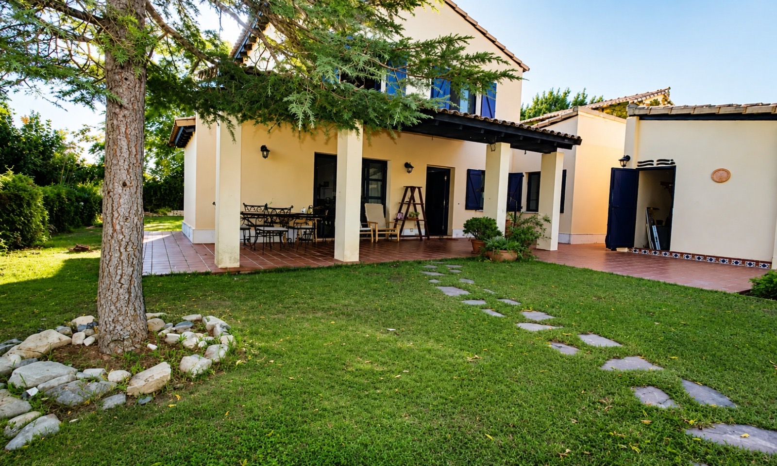 Cream-colored house with a covered patio, stone path, and a tree-shaded lawn in a sunny backyard.