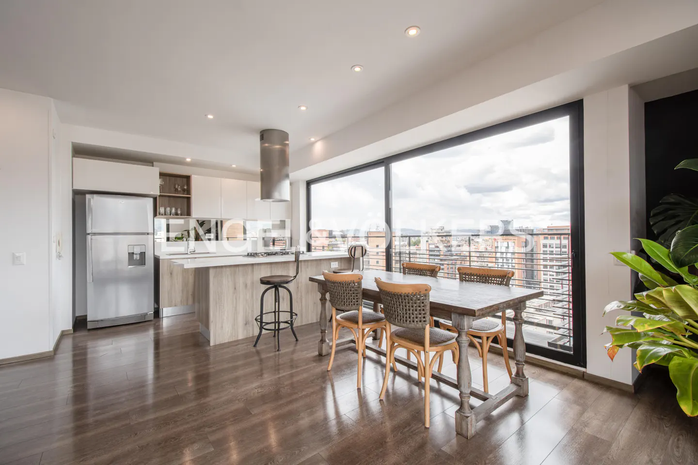 Modern kitchen with wood-look island, stainless steel fridge, and dining table by a large window overlooking the city.