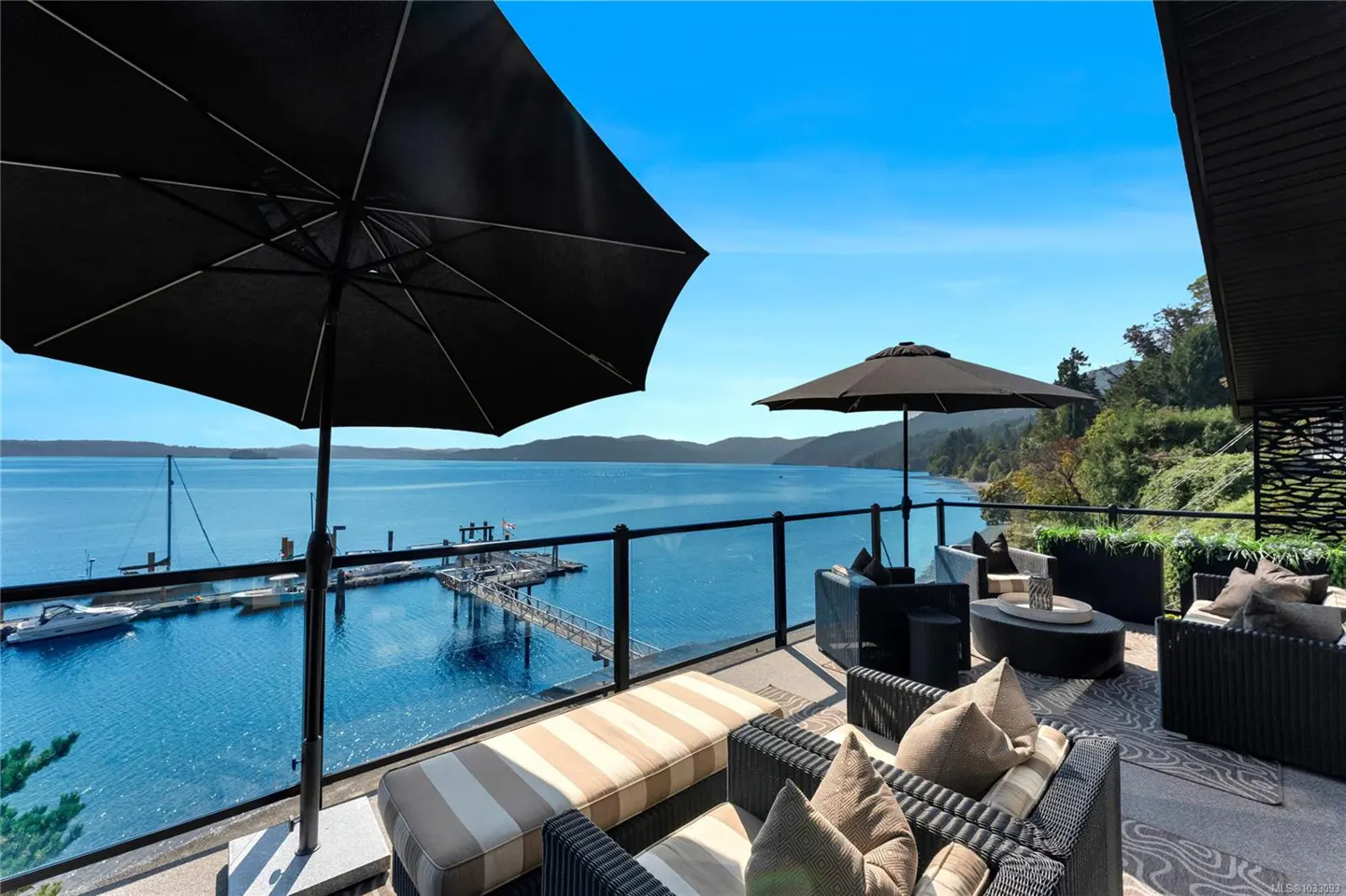 Outdoor patio with black umbrellas, wicker furniture, and a glass railing overlooking a blue ocean with a dock and boats.