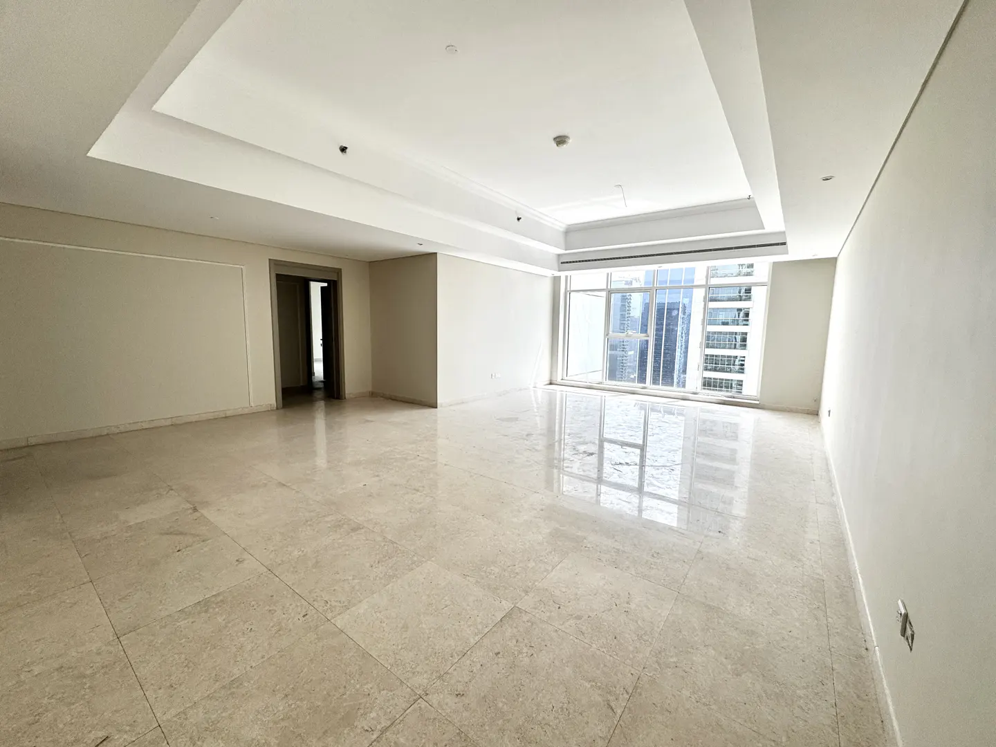 Empty, bright room with beige marble floors, white walls, and a large window showing a city skyline.