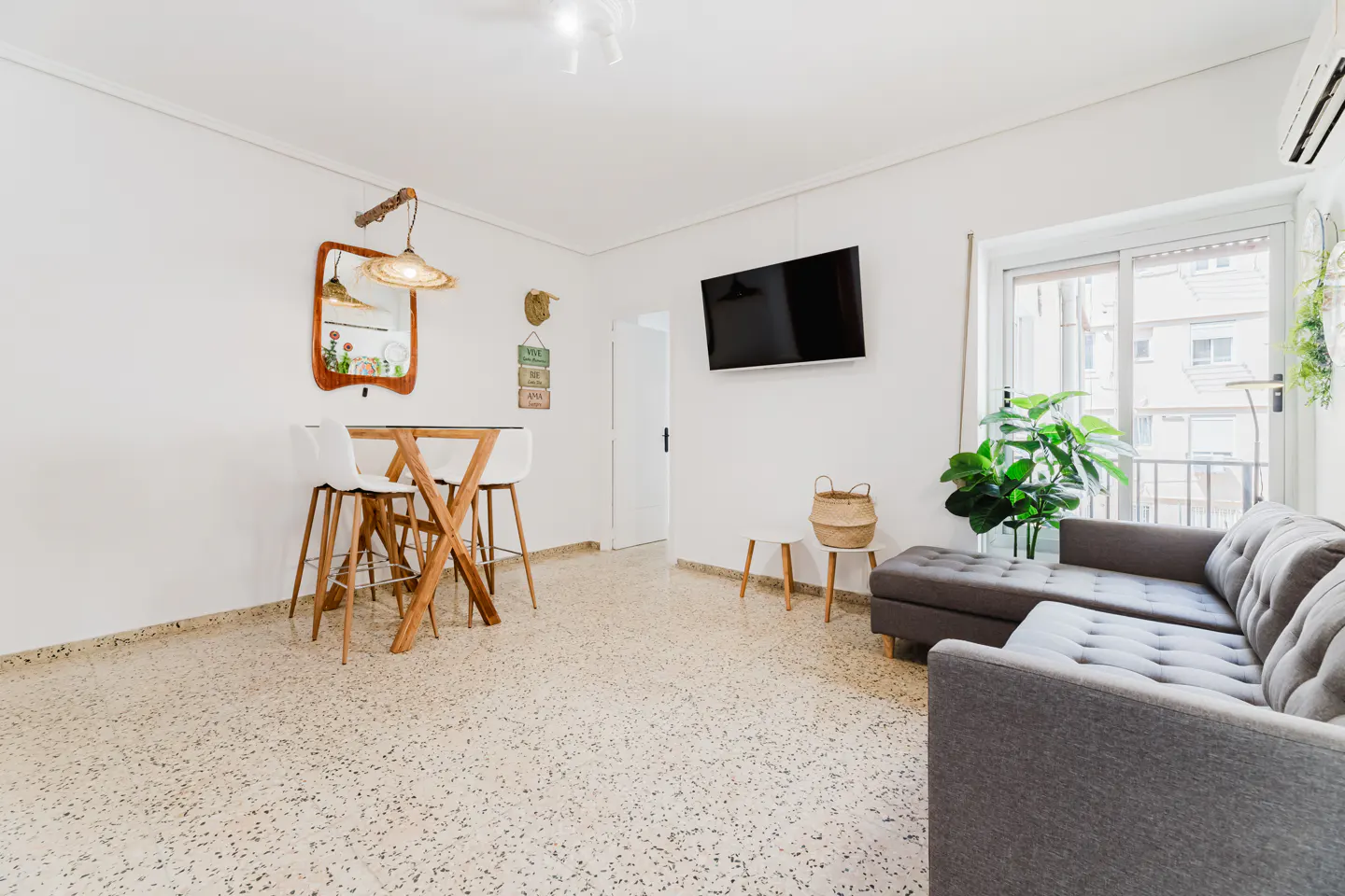 Bright living room with white walls, terrazzo floor, gray sectional sofa, TV, and wooden table with white chairs.