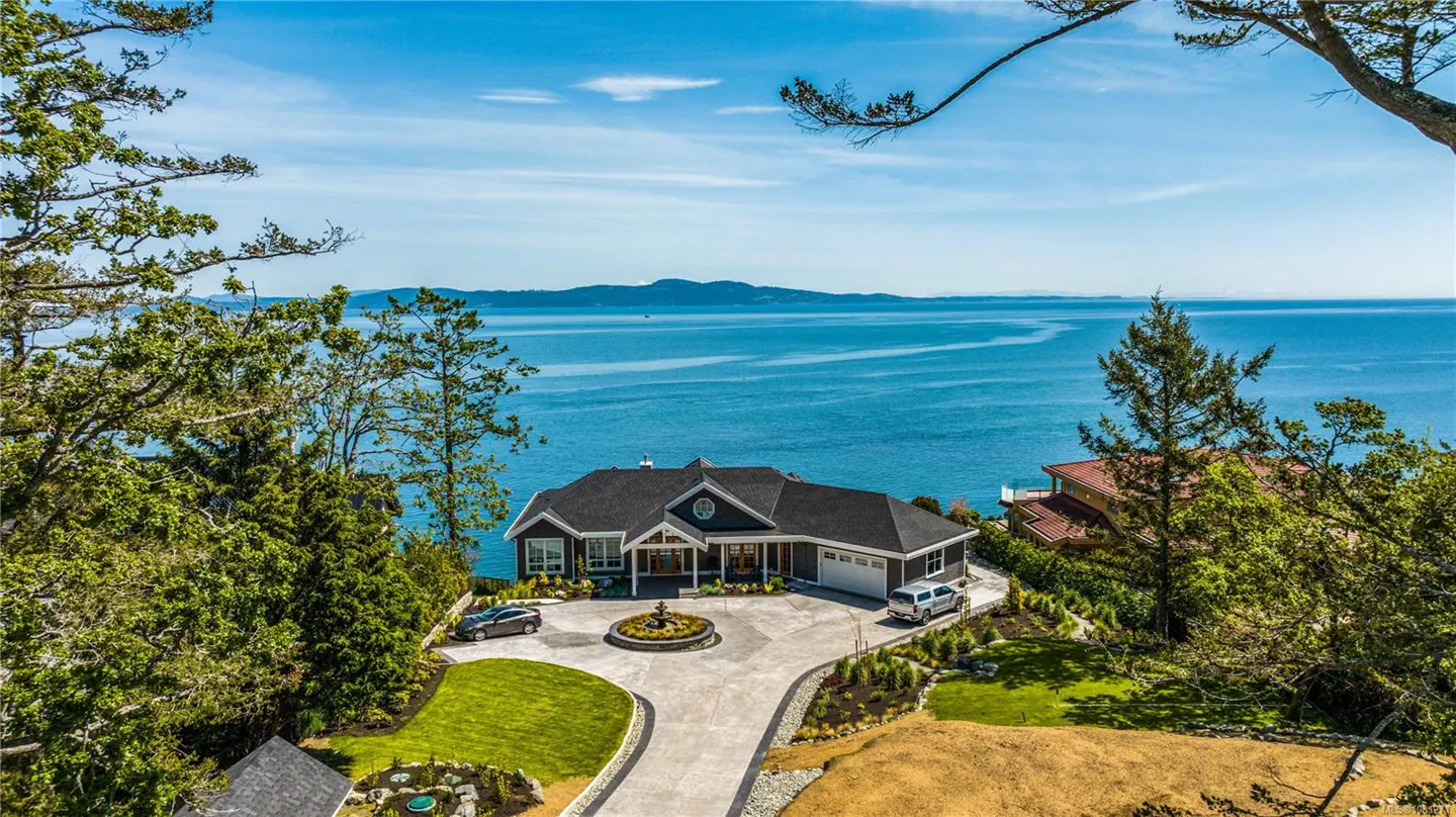 Aerial view of a dark gray house with a black roof, circular driveway, fountain, and ocean view.