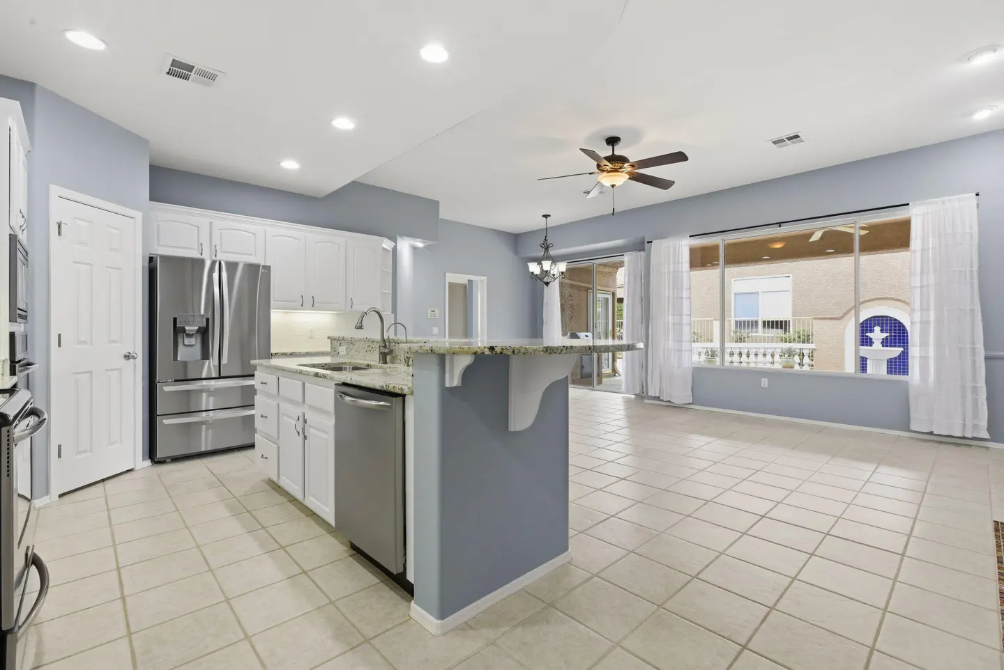 A bright, open kitchen with white cabinets, stainless steel appliances, and a granite-topped island. Large windows overlook a patio with a fountain.