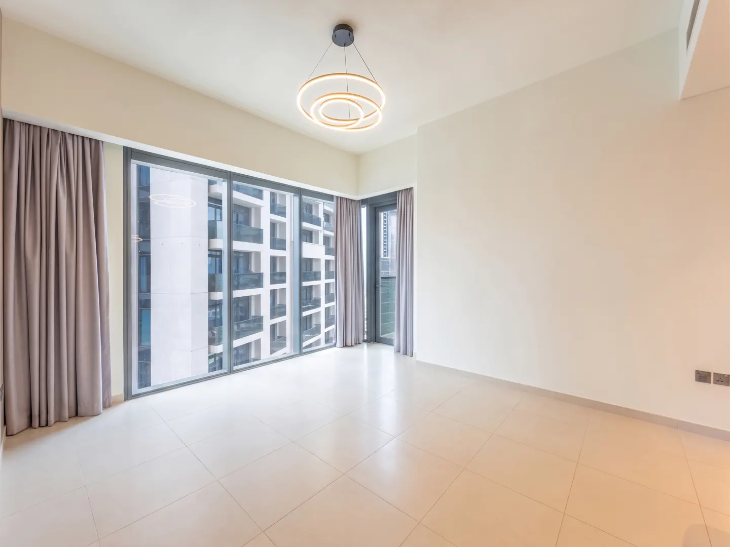 Empty room with beige tile floors, white walls, a modern circular chandelier, and floor-to-ceiling windows with grey drapes.
