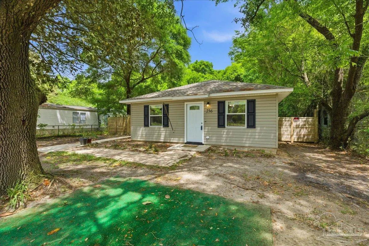 A tan, one-story house with black shutters and a white door, surrounded by green trees and a yard.