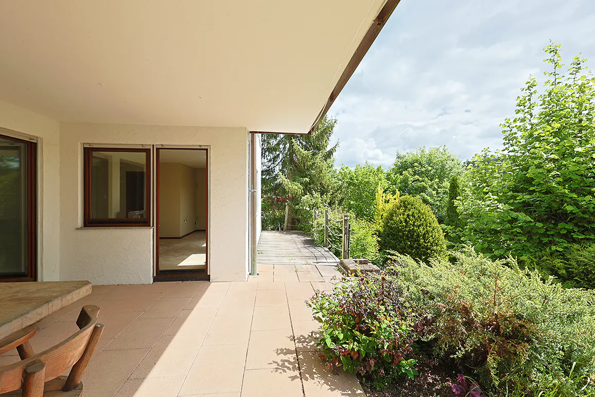 Covered patio with beige tiles, wooden chairs, and a view of a lush green garden under a cloudy sky.