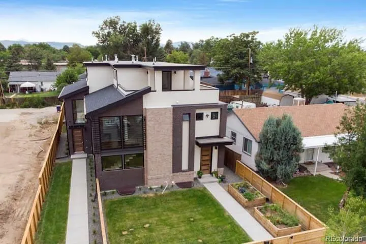Modern two-story home with brown and white exterior, large windows, and a green lawn with garden beds. Blue sky and trees in the background.