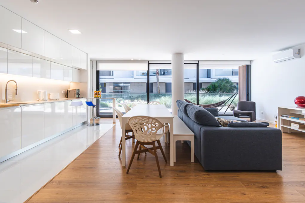 Bright, modern apartment with white kitchen, dining table, and gray sofa. Large windows overlook greenery and a hammock. Wood floors add warmth.