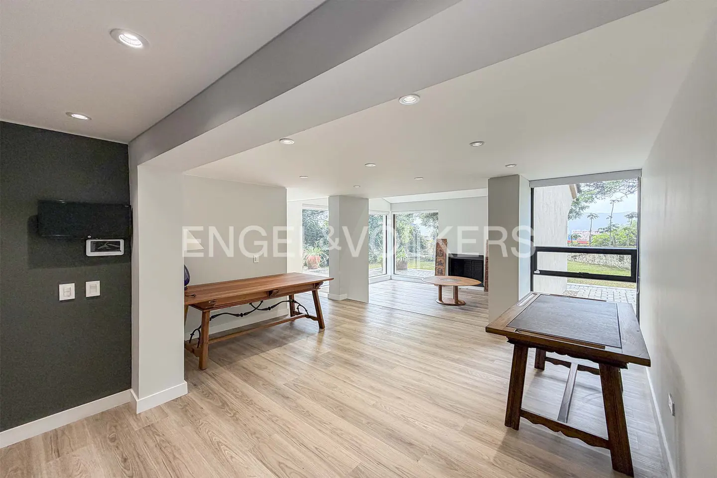 Modern open-plan room with light wood floors, two wooden tables, recessed lighting, and large windows overlooking a garden.