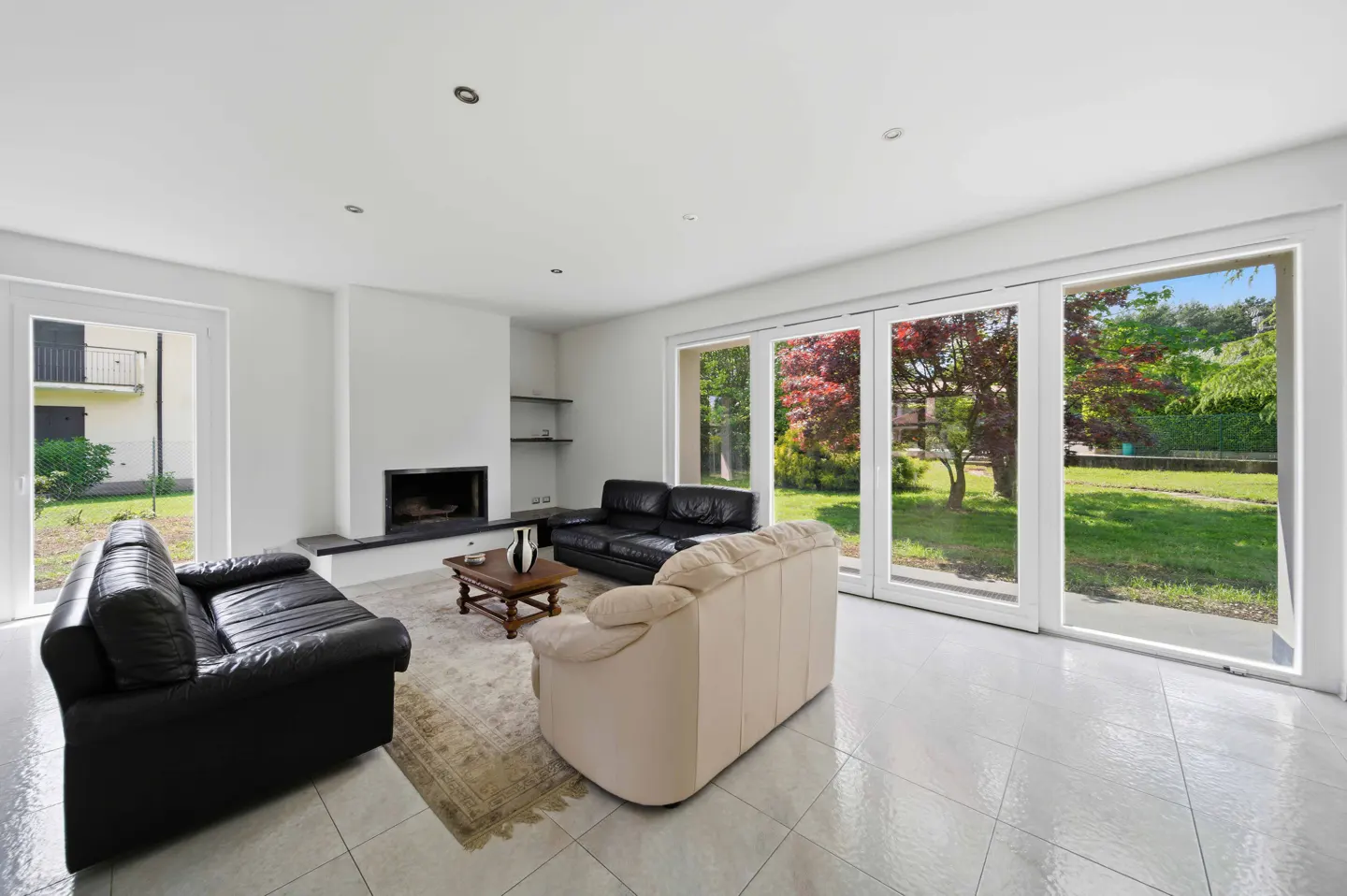 Bright living room with black and beige sofas, fireplace, and large windows overlooking a green lawn.