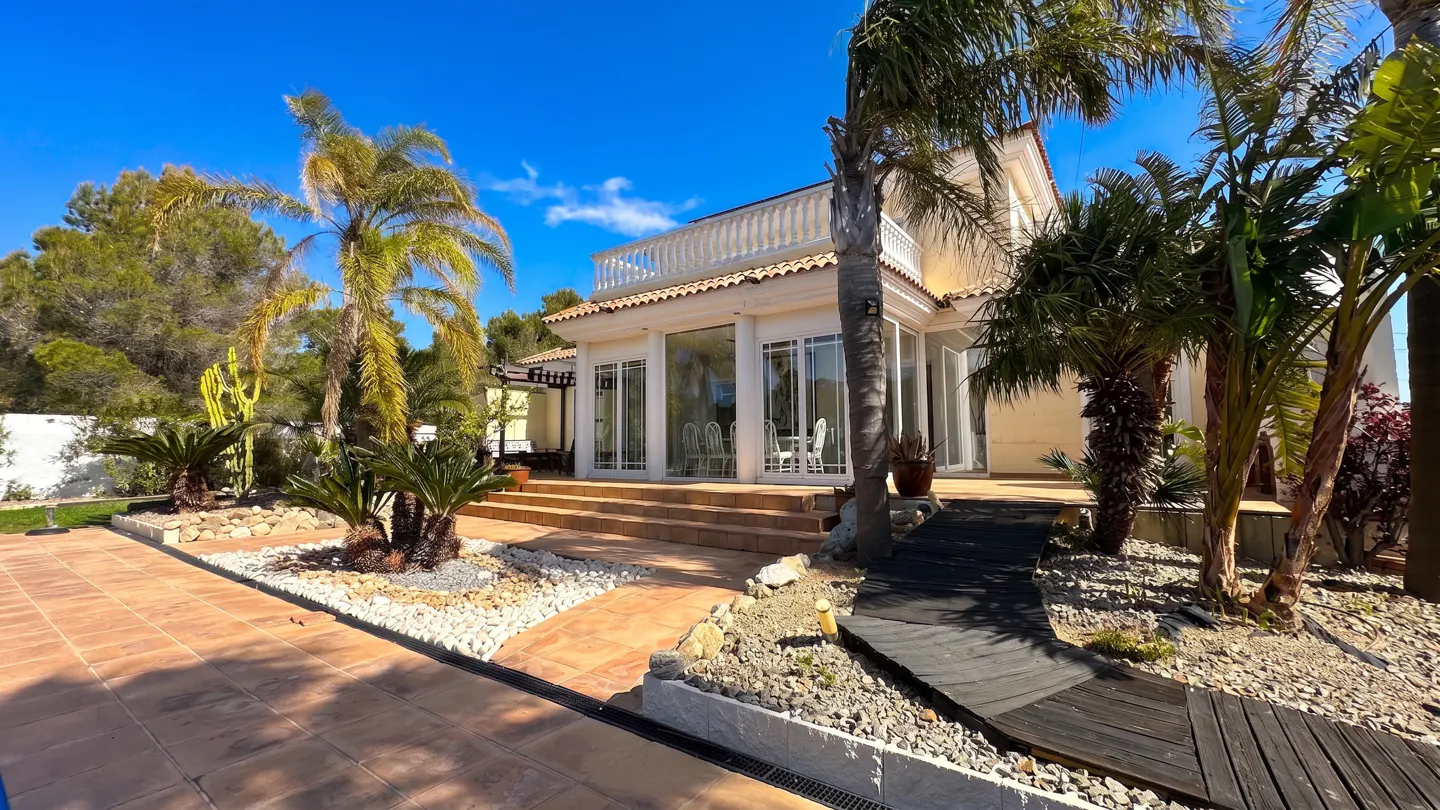 Exterior view of a tan house with a balcony, palm trees, and a wooden walkway on a sunny day.