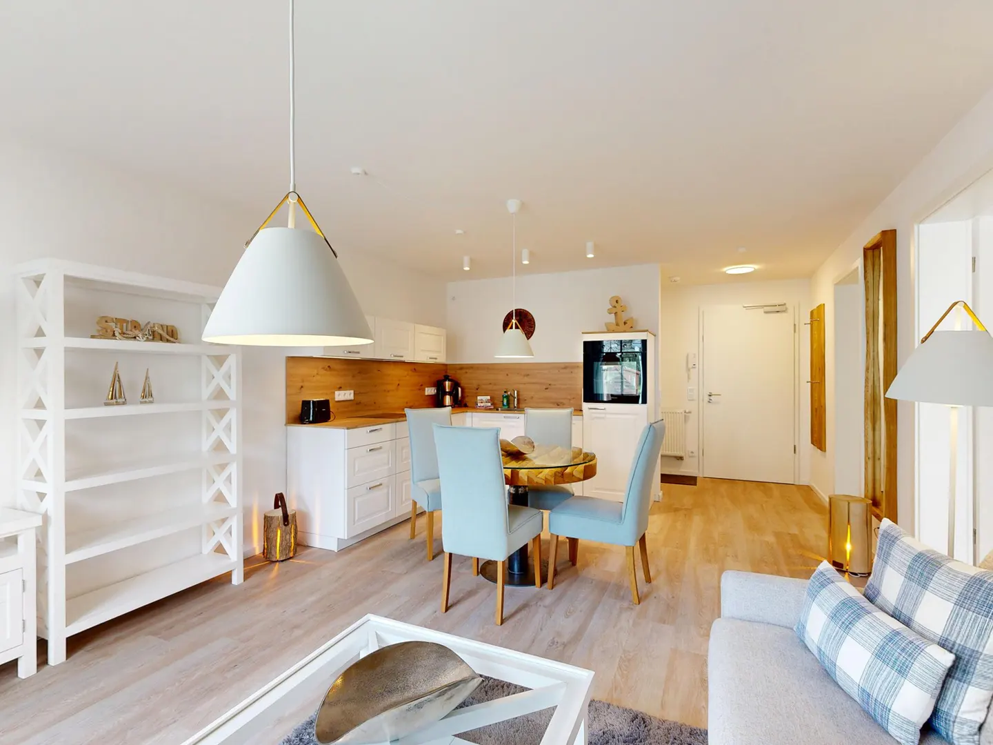 Bright, open-concept living space with white walls, wood floors, and modern furnishings. A round dining table with blue chairs sits near a white kitchen.