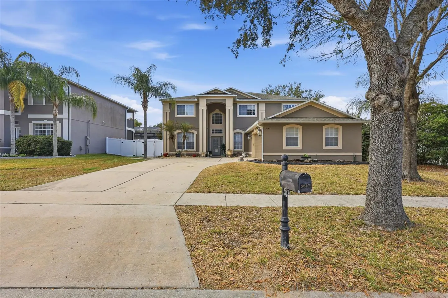 Two-story taupe house with a long driveway, palm trees, and a black mailbox on a sunny day.
