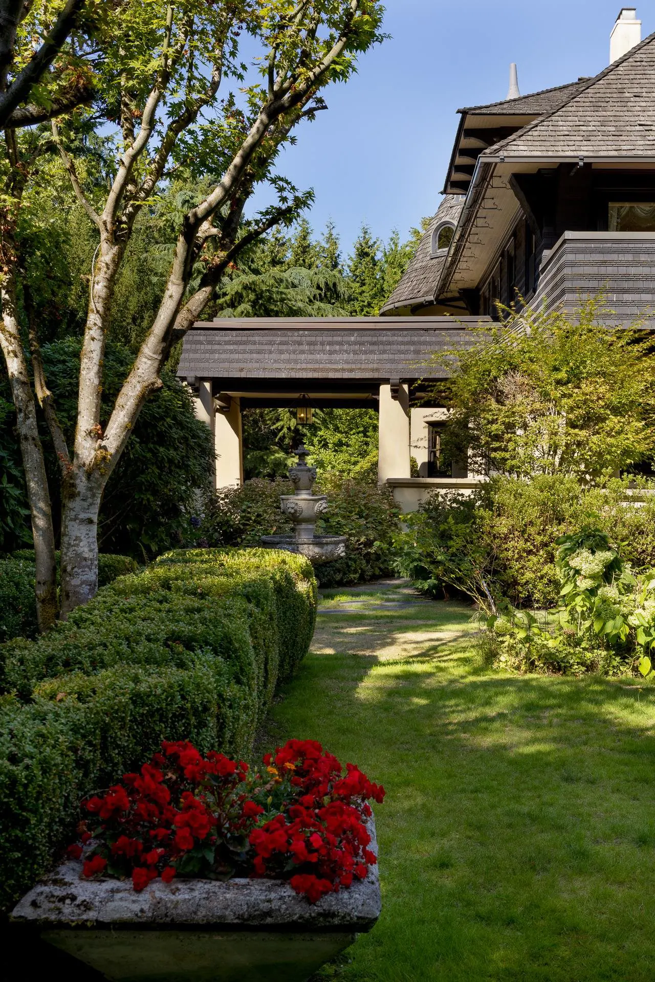 Lush green lawn and manicured hedges lead to a covered walkway connecting to a shingled house. Red flowers add a pop of color.