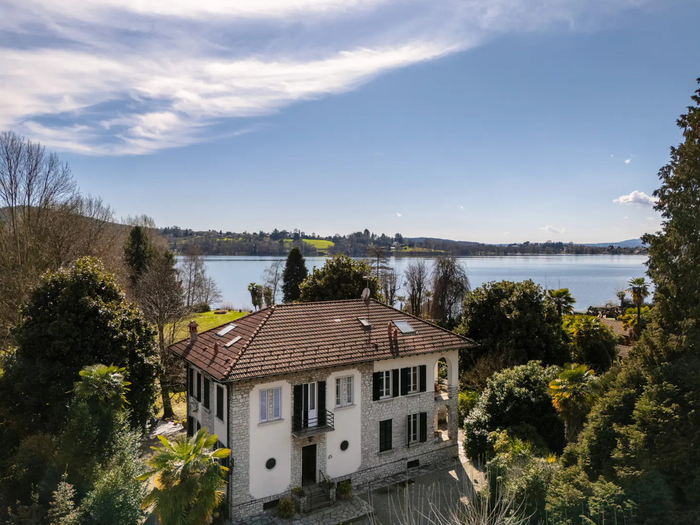 Aerial view of a white stone house with a brown tile roof, surrounded by trees, overlooking a blue lake under a blue sky.