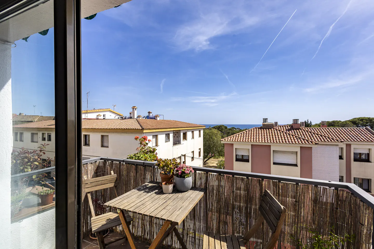 Balcony view with wooden table, chairs, and potted plants. Buildings and blue sky with clouds in the background.