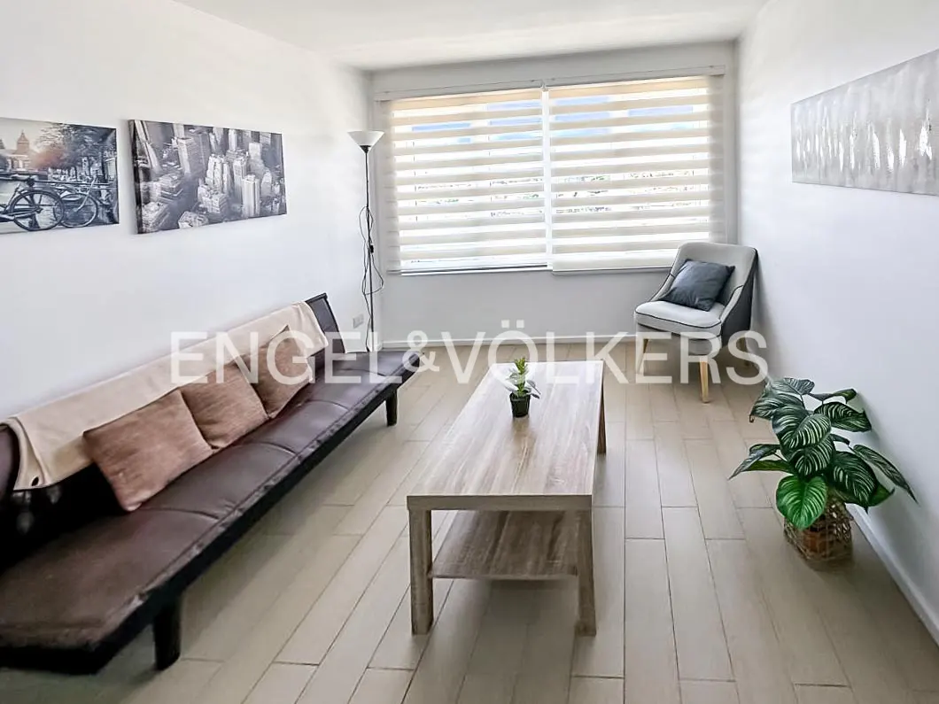 Living room with a brown futon, wood coffee table, gray chair, and artwork on white walls. A plant sits near a window with blinds.