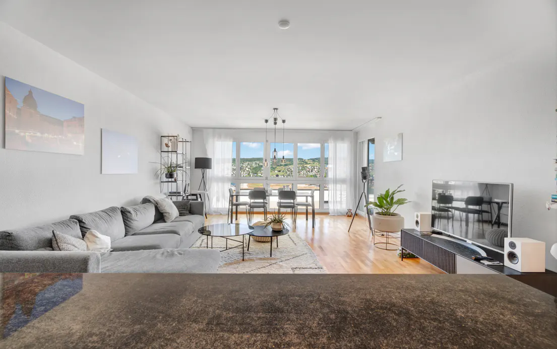 Bright living room with grey sectional sofa, round coffee tables, dining area, and large windows with a scenic view.