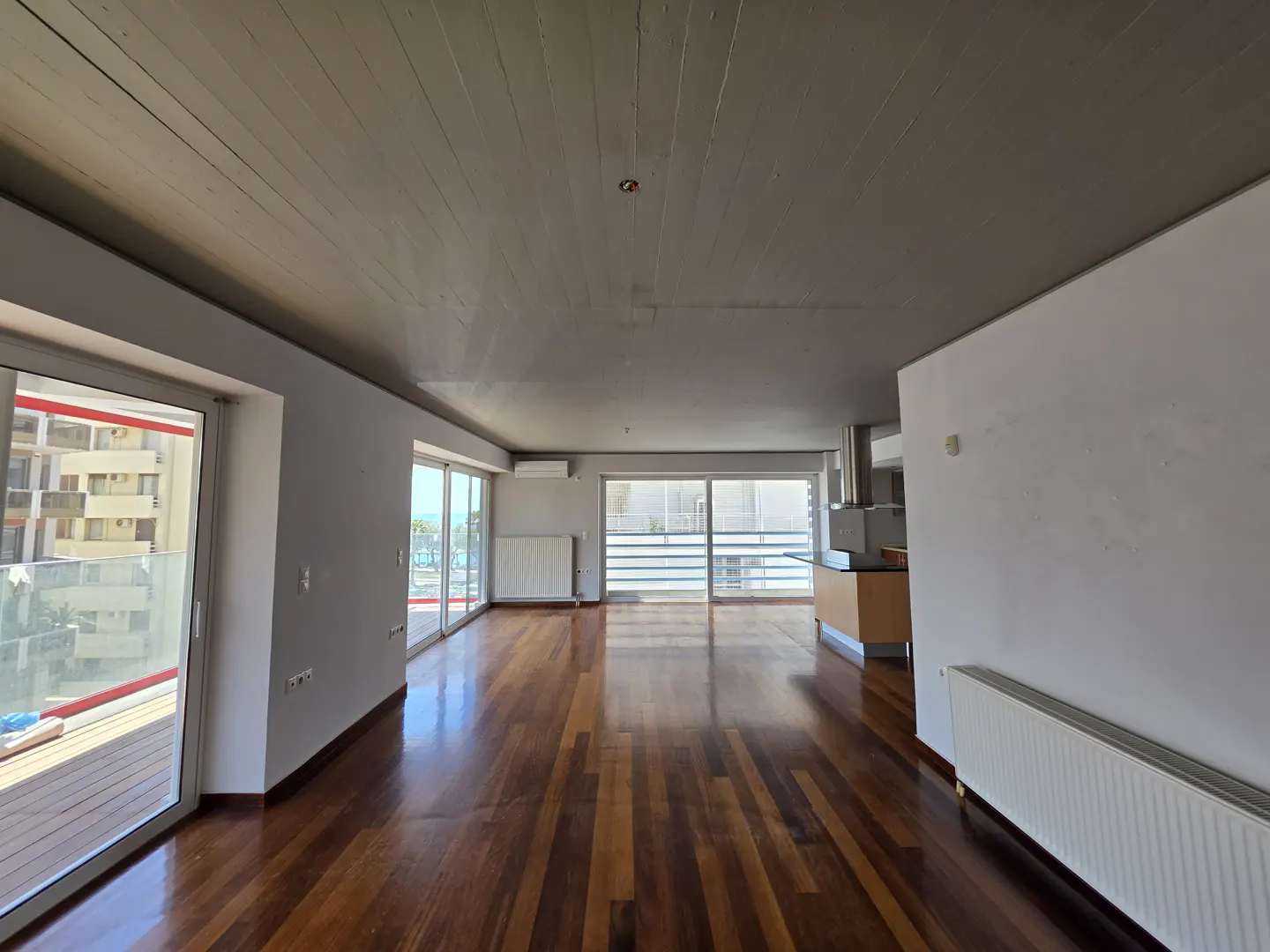 Bright, empty apartment with hardwood floors, white walls, and a gray ceiling. Balcony doors offer a view of the city and sea.