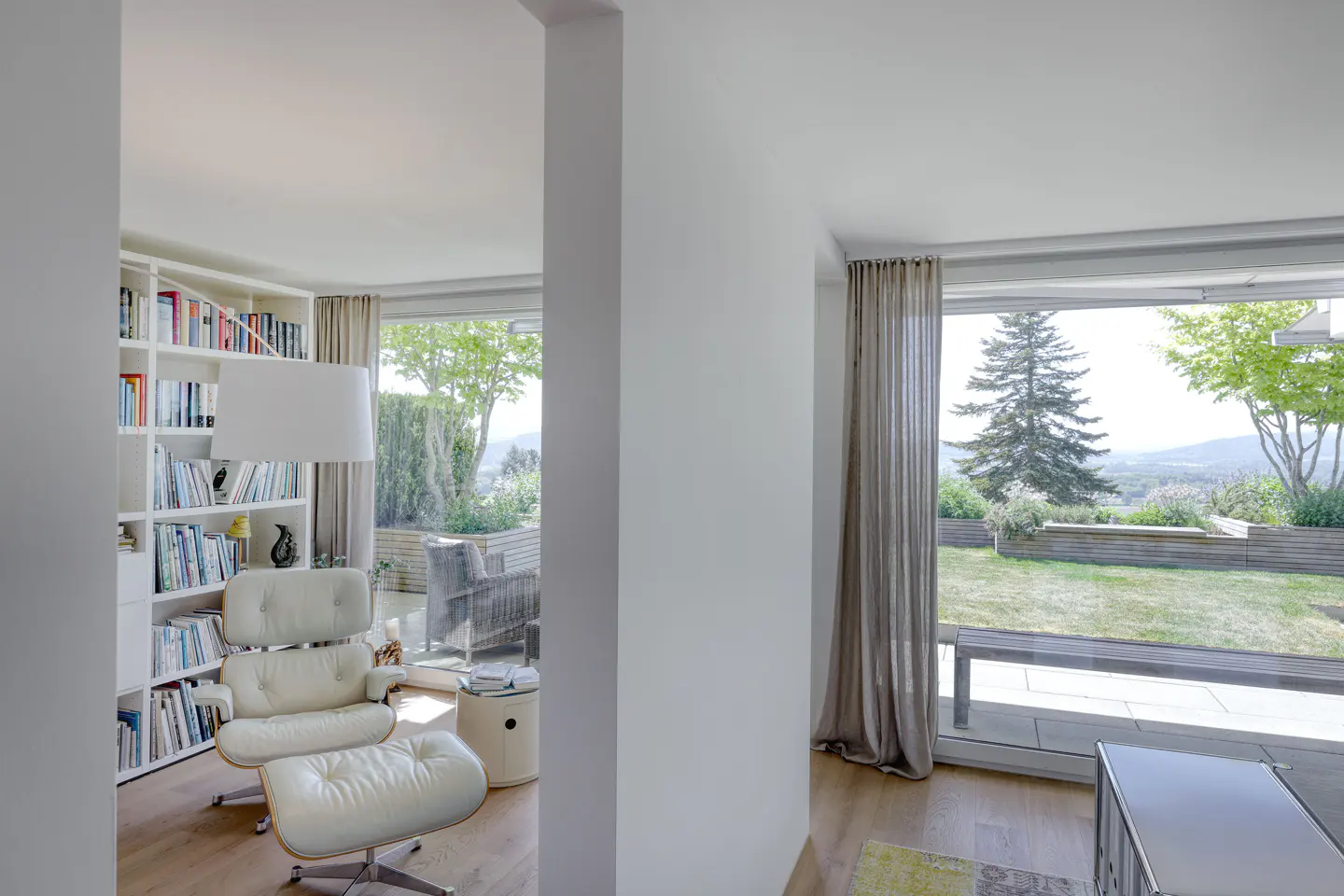 Bright living room with white walls, wood floors, and large windows overlooking a green lawn and trees. A white leather chair and bookcase are visible.