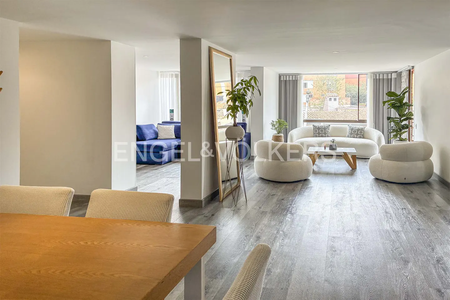 A bright, modern living room with gray wood floors, white walls, and neutral furniture. A blue sofa is visible in the background.