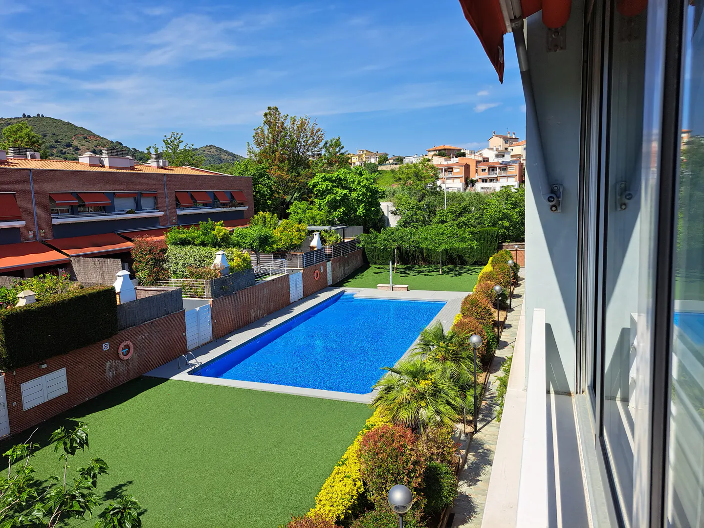 View from a window of a blue swimming pool surrounded by green grass and bushes, with buildings in the background.