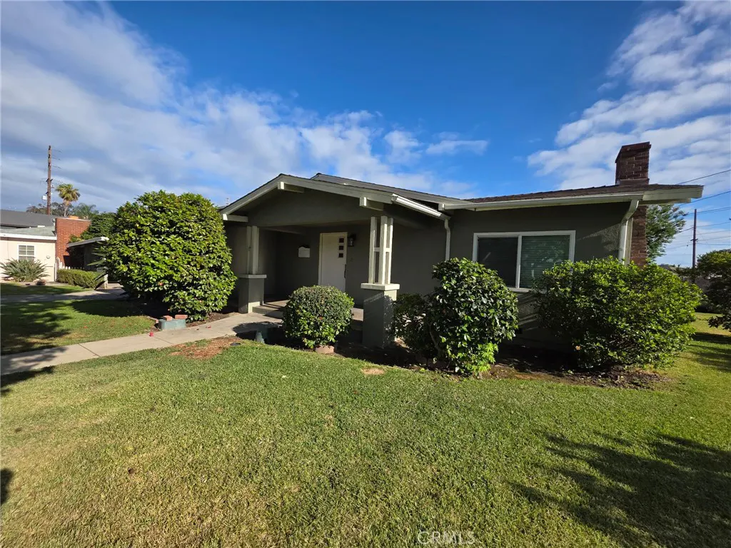 A single-story, gray house with a covered porch, white door, and green bushes under a blue sky.