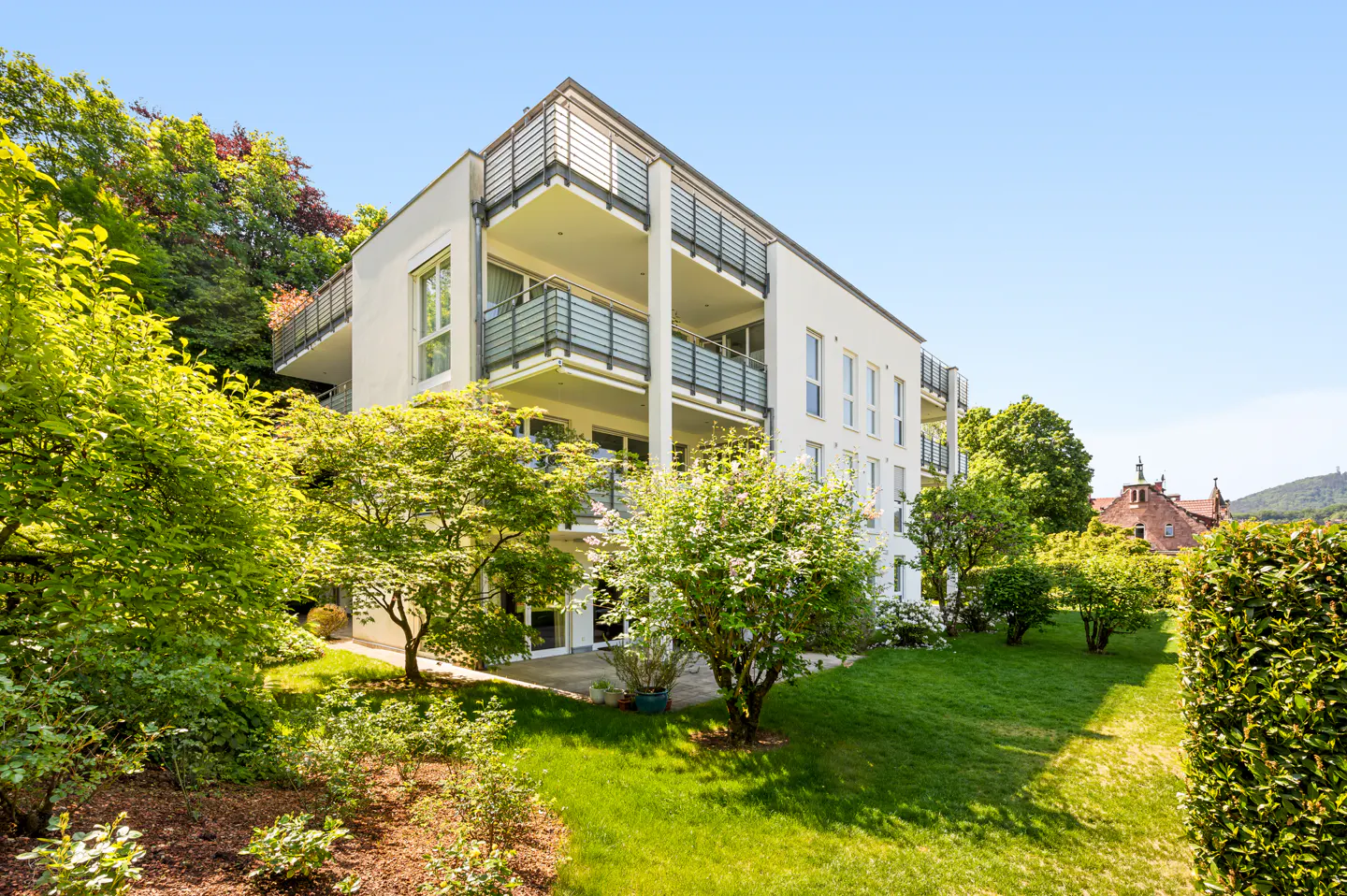 Modern white apartment building with balconies, surrounded by lush green trees and lawn on a sunny day.