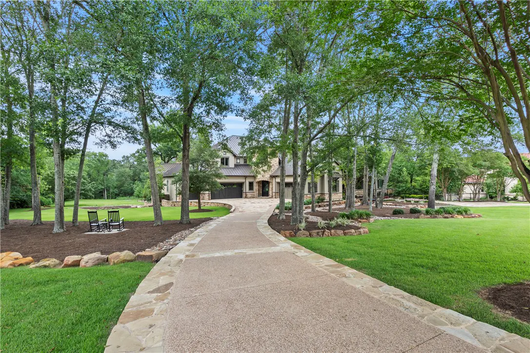 A long driveway leads to a large, two-story stone house surrounded by green trees and grass. Two black rocking chairs sit on the lawn.