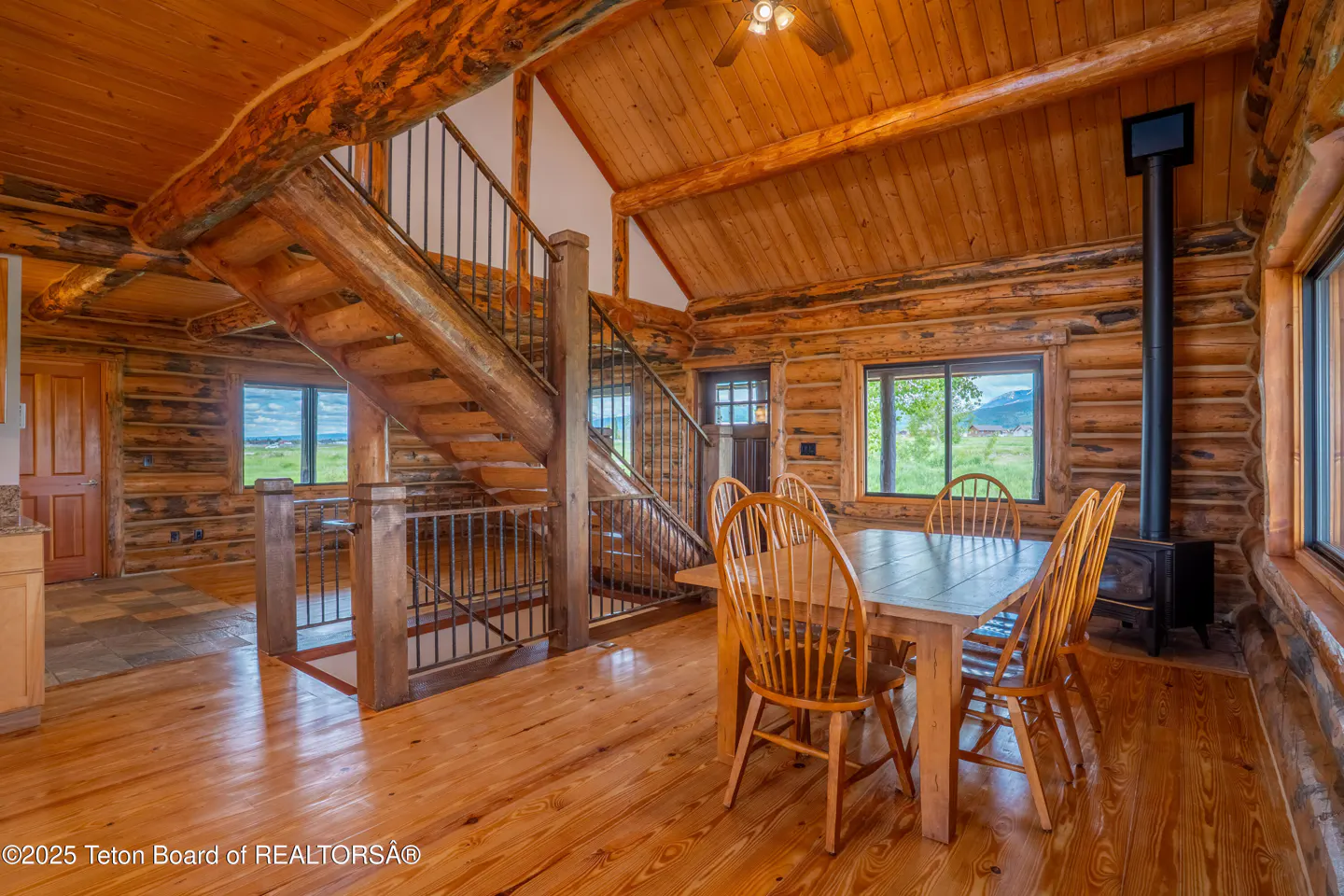 Interior of a log cabin with wood floors, a dining table with chairs, and a staircase with metal railings. Windows overlook a green field.