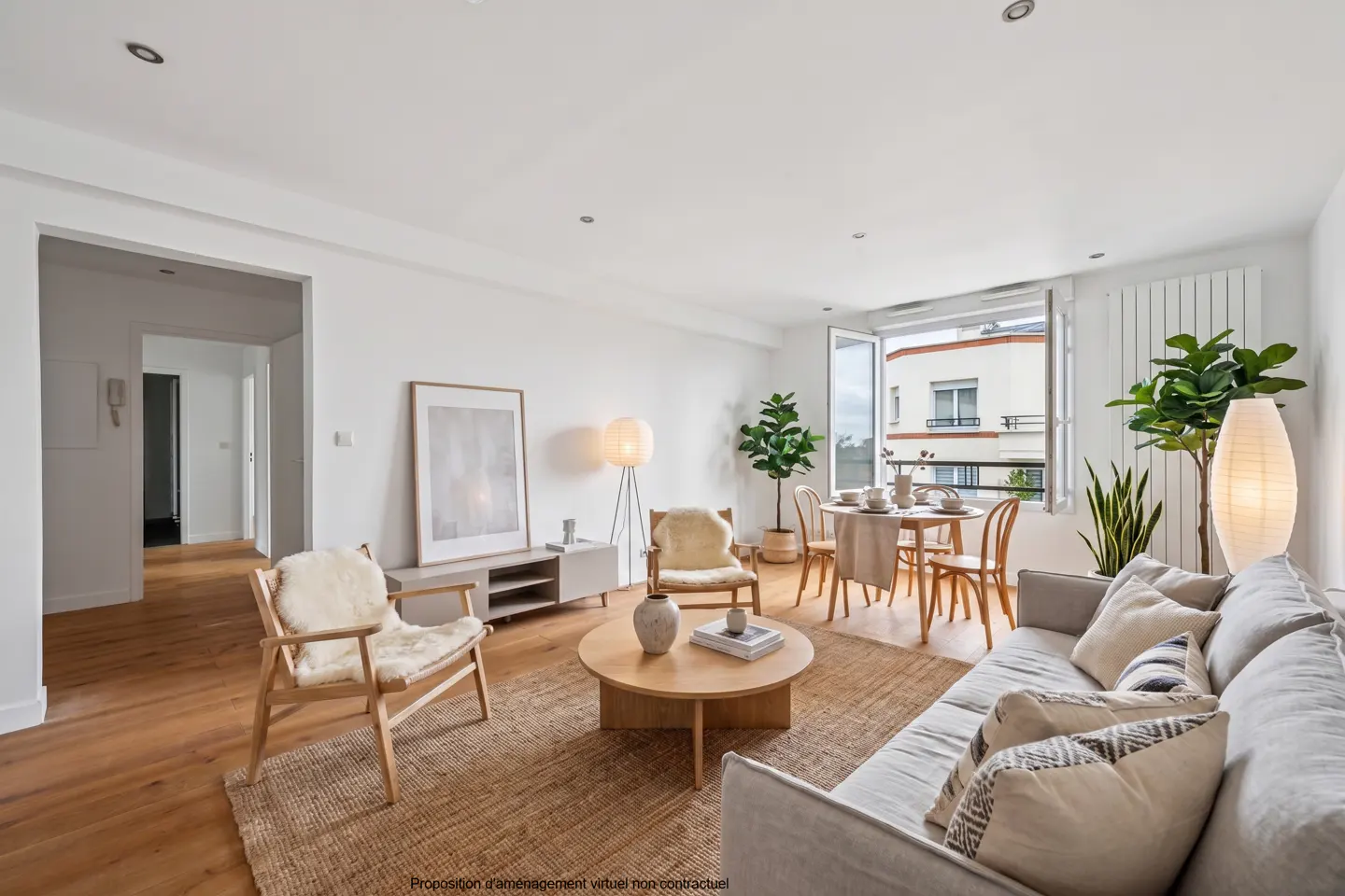 Bright living room with wood floors, a jute rug, and white walls. A gray sofa faces a round coffee table and two chairs. A dining table sits near an open window.