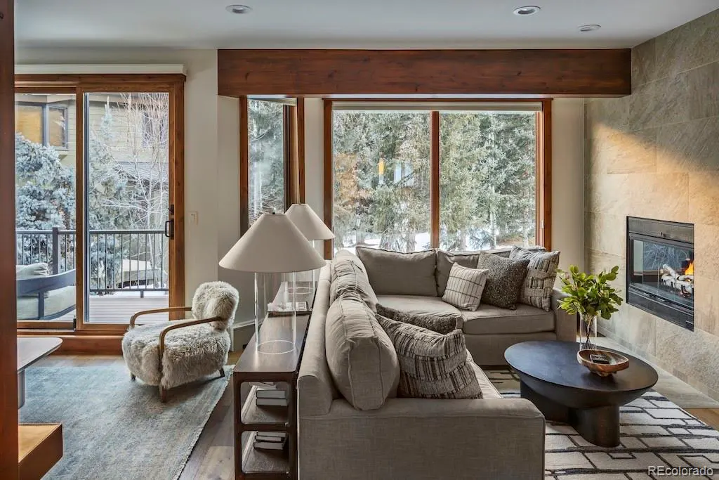 Living room with gray sectional sofa, fireplace, and large windows overlooking snow-covered trees. A furry chair sits near a sliding glass door.