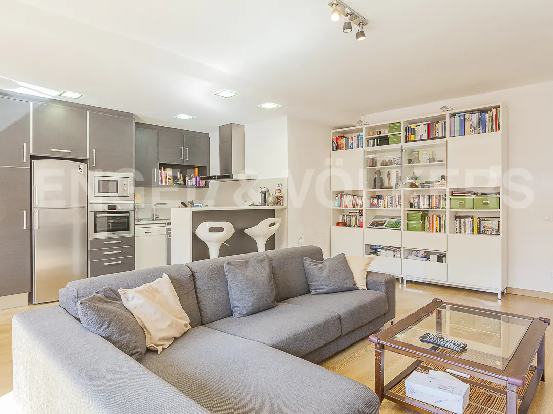Open-concept living space with gray sectional sofa, wood coffee table, and white bookcase. Kitchen with gray cabinets and bar stools in background.