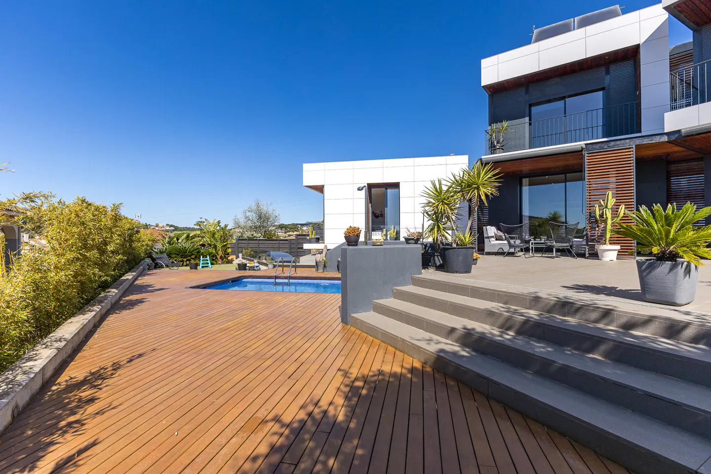 Modern home exterior with a wooden deck, pool, and patio furniture under a clear blue sky.