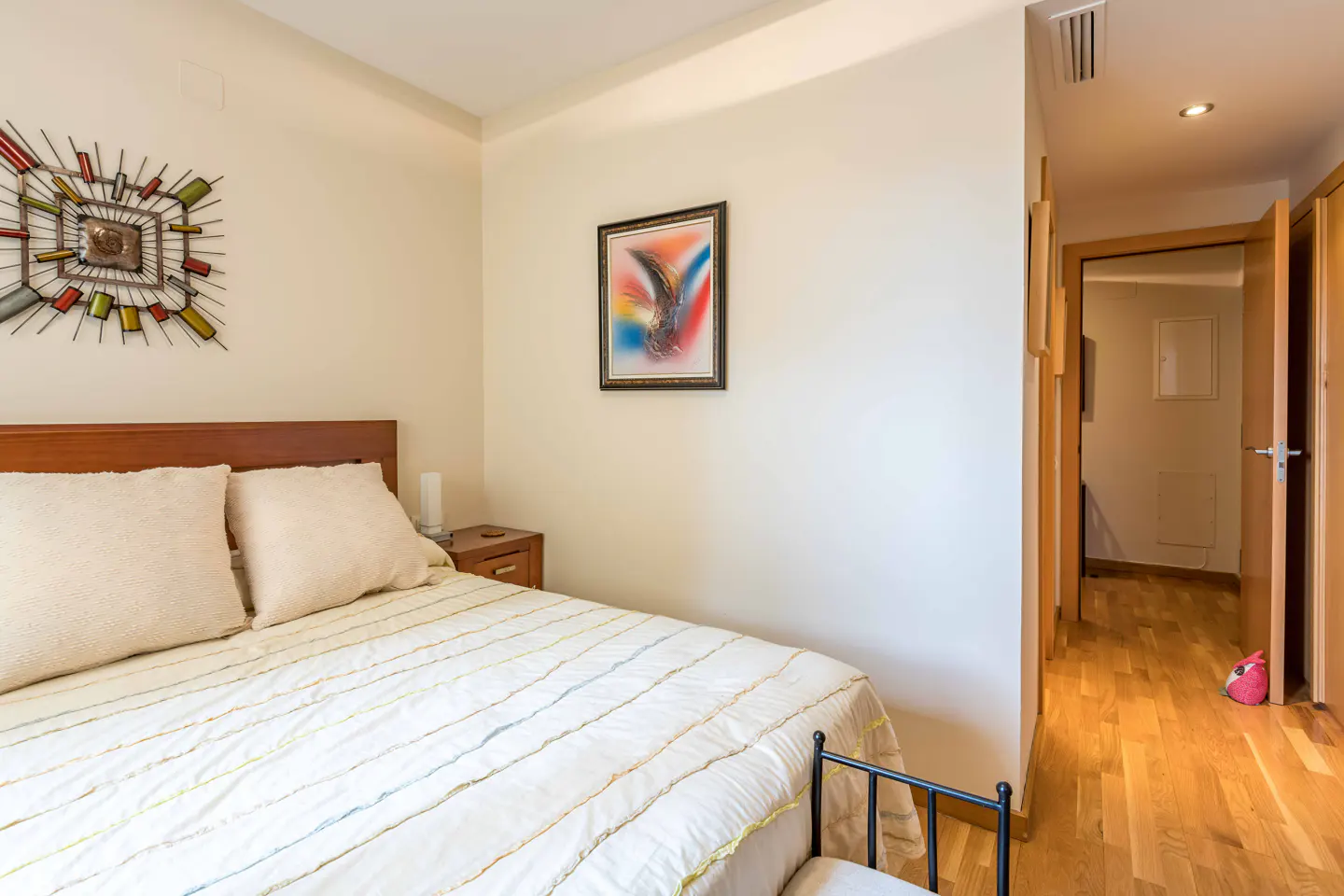Bedroom with a wooden bed frame, white bedding, and a colorful metal wall decoration. A hallway with wood floors is visible.