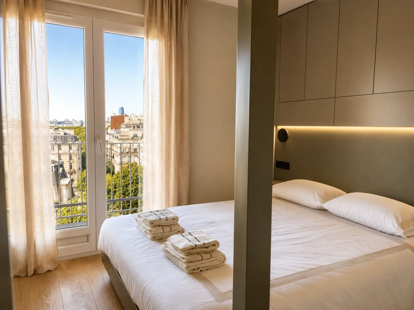 Bedroom with white linens, stacked towels, and a view of city buildings through a window with beige curtains.