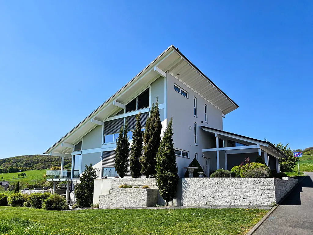 Modern gray house with a white roof and trim, set on a green lawn with trees and a stone wall under a clear blue sky.