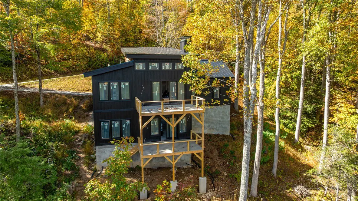 Modern black house with a metal roof and two-story wooden deck, surrounded by trees with autumn foliage.
