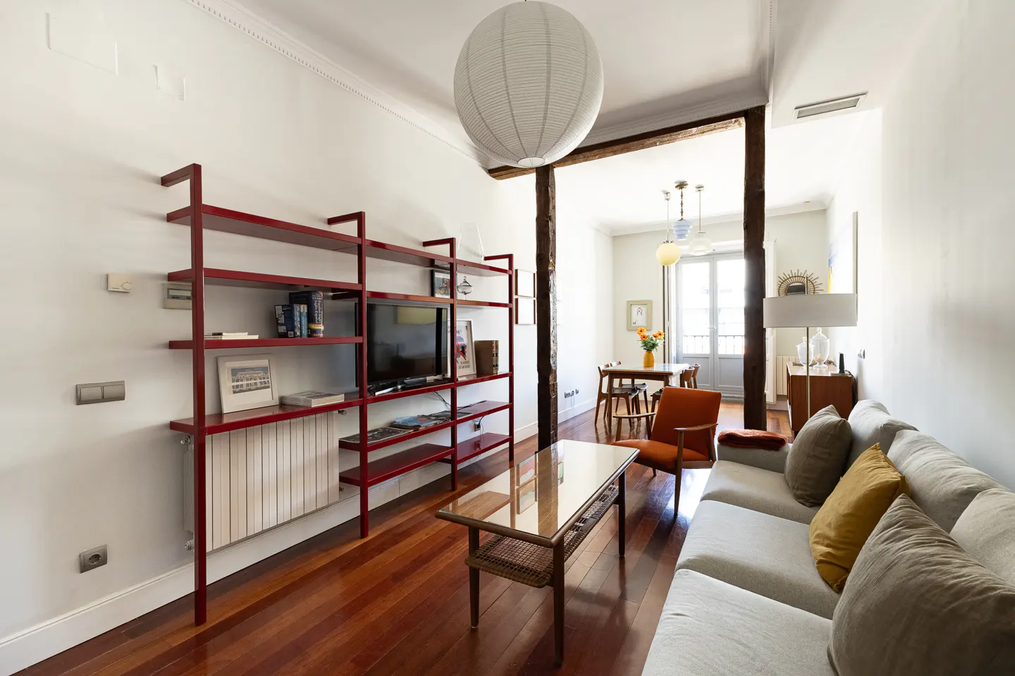 Modern living room with a red metal shelving unit, grey sofa, orange armchair, glass coffee table, and hardwood floors.
