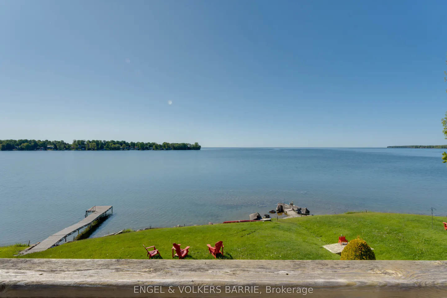 Waterfront view with a dock, red Adirondack chairs on green grass, and a clear blue sky.
