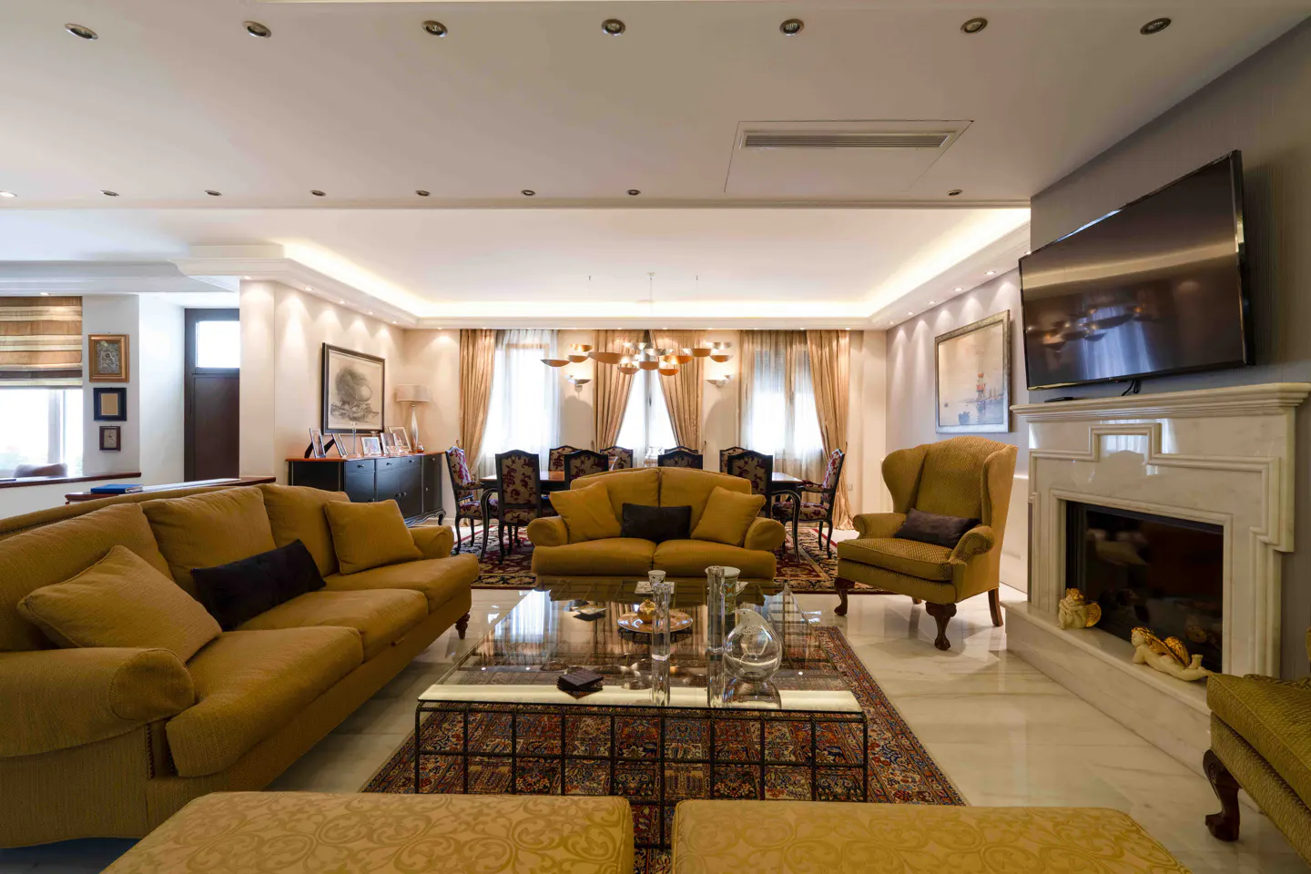 A spacious living room with yellow sofas, a glass coffee table, and a dining area in the background. A TV is mounted above a marble fireplace.