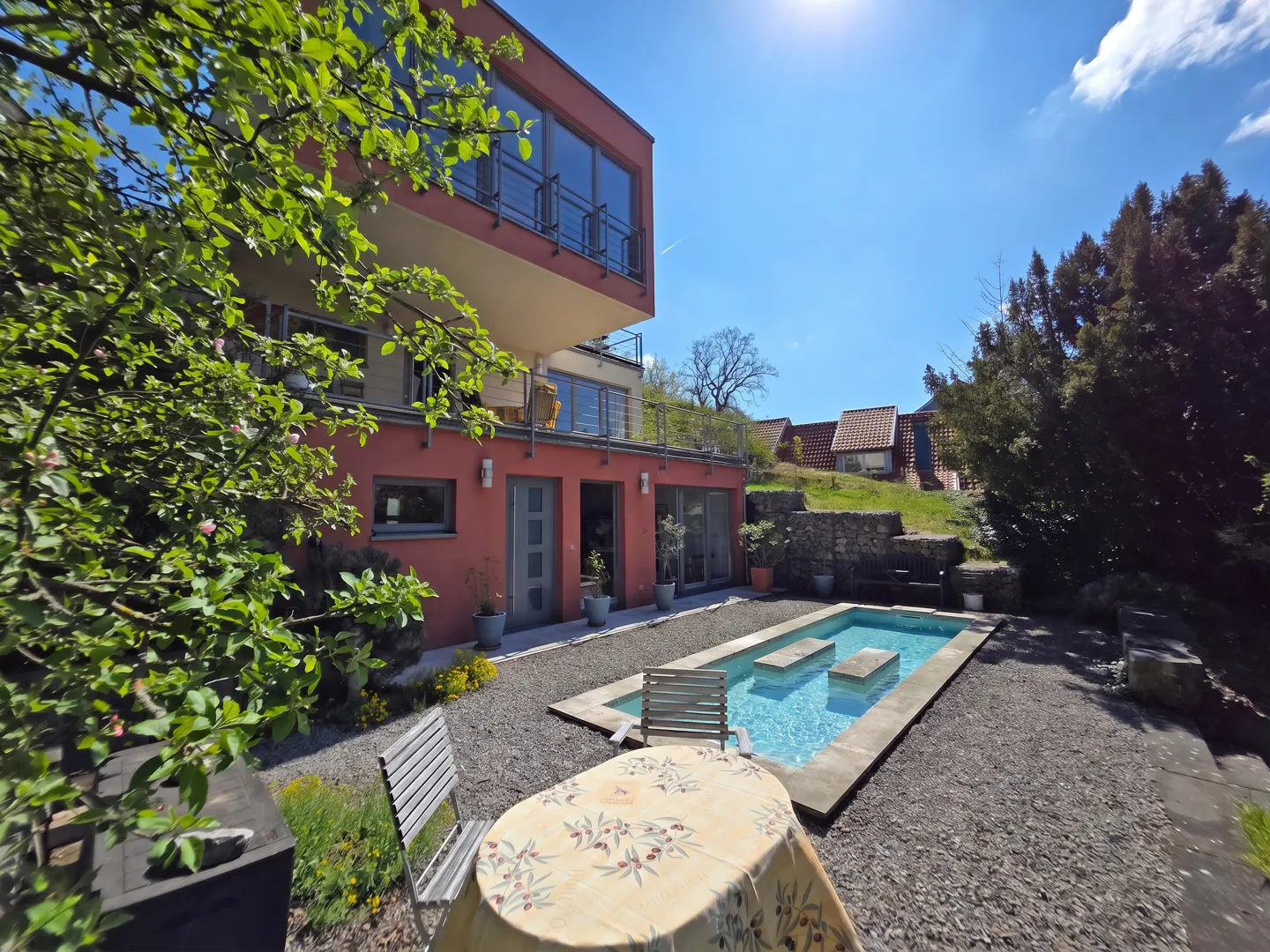 Backyard view of a modern, two-story red house with a pool, table, and chairs on a sunny day.
