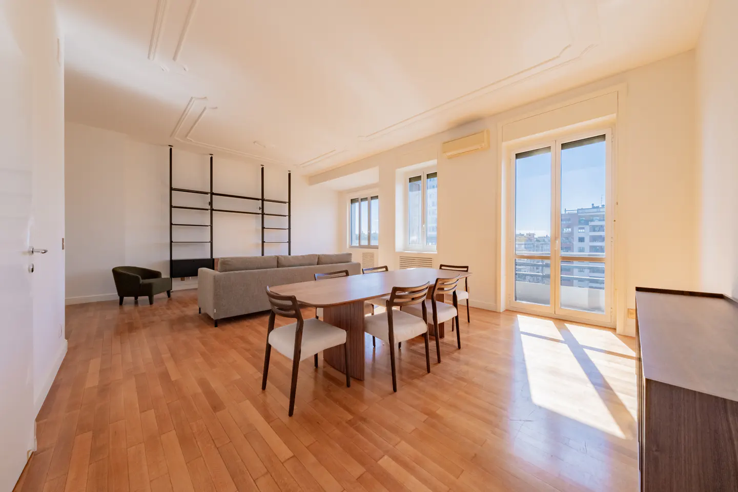 Bright living room with hardwood floors, a dining table with six chairs, a gray sofa, and a black bookshelf. Large windows offer city views.