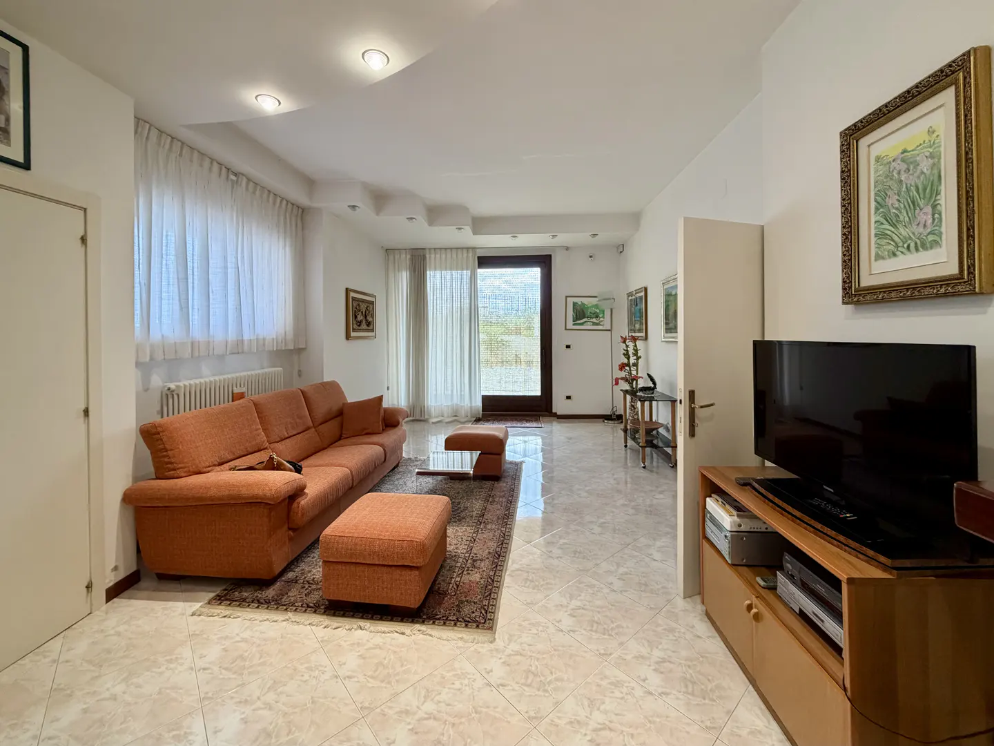 A bright living room with an orange sofa, ottoman, and a patterned rug on a tiled floor. A TV sits on a wooden stand. Art adorns the white walls.
